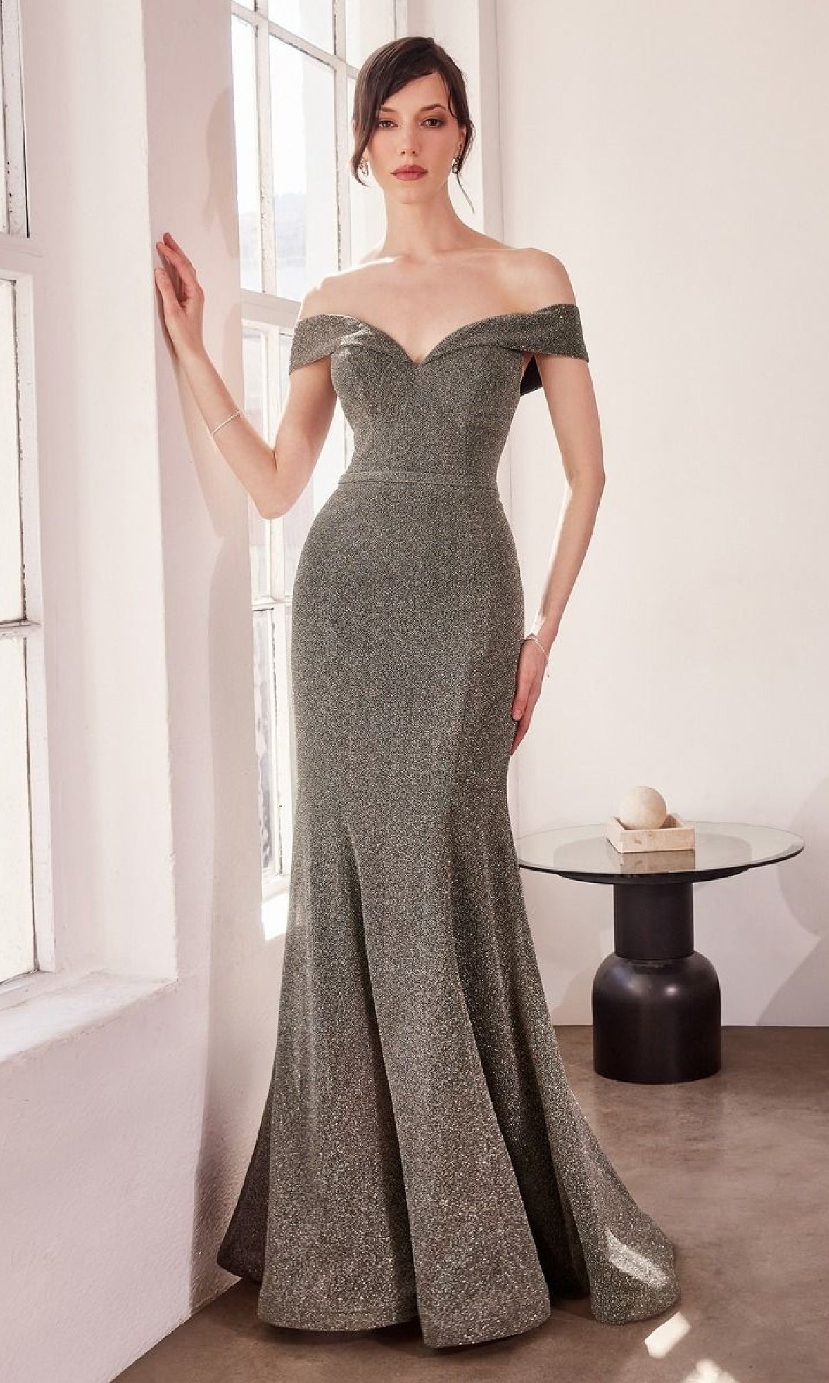 A woman stands indoors by a window wearing the Off-Shoulder Sweetheart Prom Gown: Amoris T602, a fitted metallic silver evening dress with a flared skirt. Beside her is a small decor table.